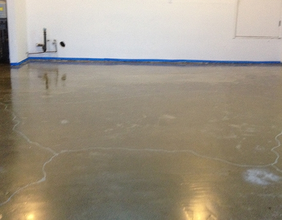 An empty room with smooth, polished concrete flooring, ideal for decorative concrete coatings for garage spaces. The floor reflects light, and blue painter’s tape lines the base of the white wall. Exposed pipes and a door are visible in the background.