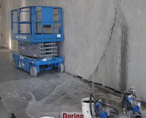 A blue scissor lift is parked beside a concrete wall inside a building. Buckets, hoses, and equipment for polyurethane grout injection are on the floor, with netting spread out. The word "During" appears in red text.
