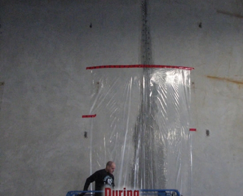 A person stands on a blue lift beside a wall, partially surrounded by a tall, transparent plastic sheet labeled "During," with a vertical streak—likely from polyurethane grout injection—visible behind the plastic.
