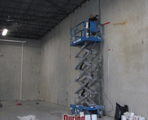A person on a blue scissor lift works near the top of a high, bare concrete wall inside an industrial building, possibly preparing for polyurethane grout injection. Paint cans and supplies are scattered on the floor. The word "During" appears at the bottom.