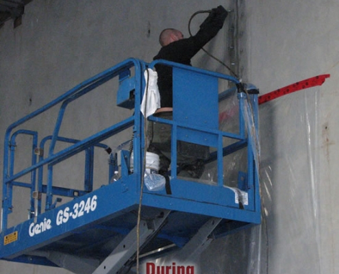 A person stands on a blue Genie GS-3246 scissor lift, working on a tall wall with tools. The word "During" is written below, indicating polyurethane grout injection is in progress.