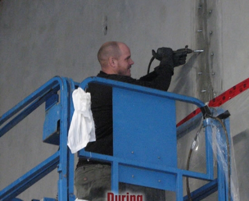 A person on a blue lift uses a power tool to repair or work on a concrete wall, possibly preparing for polyurethane grout injection. The wall has visible bolts or anchors, and the word "During" is overlaid on the image.