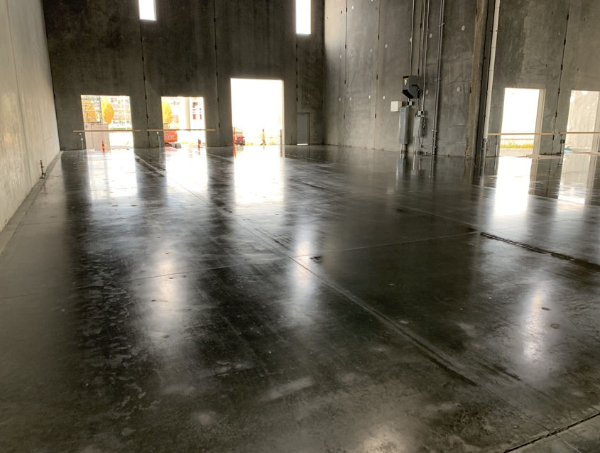 A smooth, polished warehouse concrete floor inside a large, empty industrial building with high ceilings and unfinished walls. Natural light enters through expansive door and window openings.