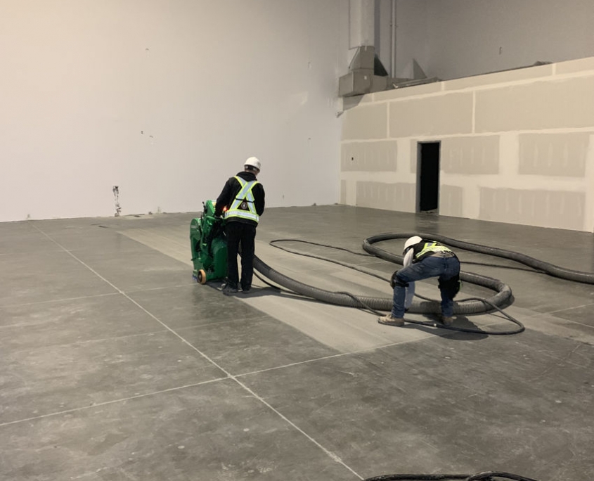 IMG_1628 Two construction workers wearing safety gear operate machinery and large hoses on the floor of a spacious, unfinished building, focusing on warehouse concrete floor repair amid bare walls and visible utility ducts.