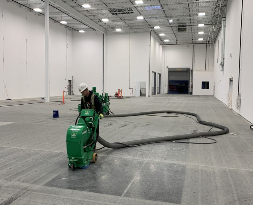 IMG_2636 A worker wearing a hard hat operates a green floor grinding machine in a large, empty warehouse, preparing the concrete floor for an industrial epoxy coating. Another similar machine is visible in the background.