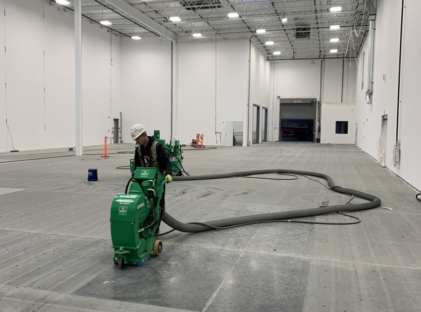 IMG_2636 A worker wearing a hard hat operates a green floor grinding machine in a large, empty warehouse, preparing the concrete floor for an industrial epoxy coating. Another similar machine is visible in the background.
