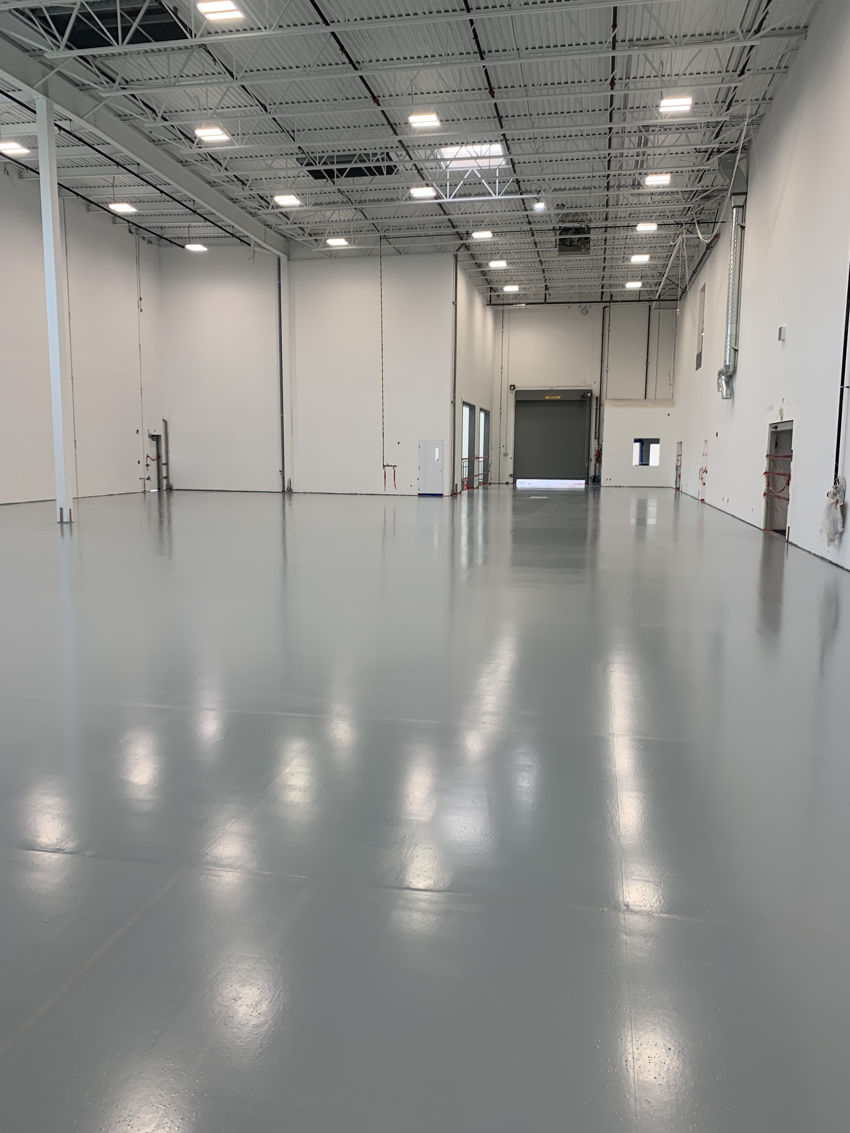 IMG_2673 A spacious, empty industrial warehouse with polished gray floors recently enhanced by warehouse concrete floor repair, high ceilings, white walls, and large overhead lights. Multiple loading dock doors and a few wall fixtures are visible along the sides.