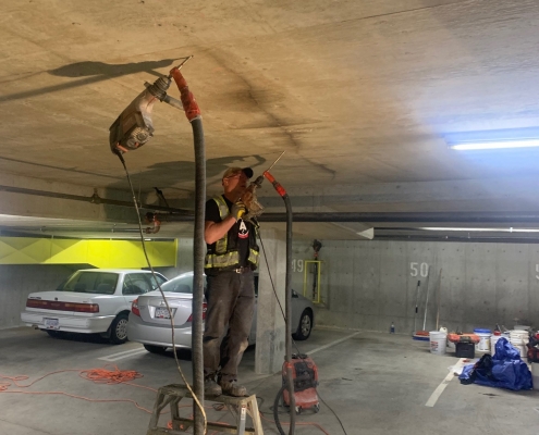 A worker in a safety vest stands on a ladder in a parking garage, using power tools for polyurethane grout injection on the ceiling. Cords and equipment are scattered on the ground, with parked cars seen in the background.