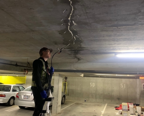 A person stands on a ladder in a parking garage, repairing a large crack in the concrete ceiling using tools and polyurethane grout injection. A blue tarp covers the floor, while cars are parked in the background.