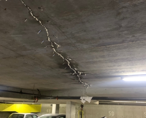 A concrete parking garage ceiling with visible cracks repaired using polyurethane grout injection; several syringes or nozzles protrude from the repair line. Cars are parked below, and a ladder is set up near the repair area.