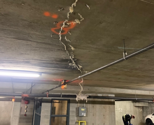 Ceiling of an underground parking garage shows a long crack and peeling material, suggesting the need for polyurethane grout injection. Spray-painted orange markings, pipes, and stacked bags and containers are visible near parking spaces.