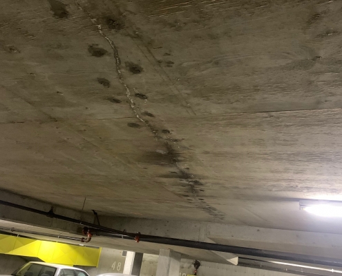 A concrete parking garage ceiling with visible water stains and cracks, suggesting the need for polyurethane grout injection. Several cars, including a black and white sedan, are parked below near yellow-marked spots numbered 48 and 49.