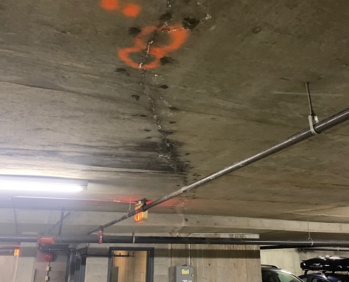 A concrete ceiling in a parking garage with a water stain and crack, marked with orange spray paint, possibly requiring polyurethane grout injection. Cars are parked nearby, and a small utility room is below the damaged area.