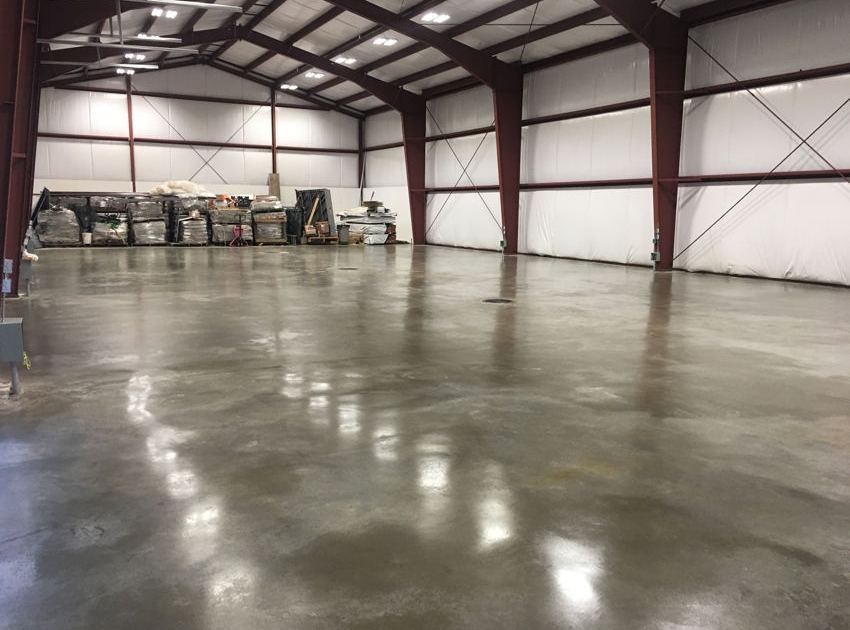 A spacious, empty warehouse with a shiny, polished concrete floor showcases recent warehouse concrete floor repair, enhanced by red steel beams and white insulated walls. Stacks of materials and equipment are stored along the back wall.