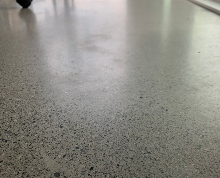 polish-concrete-1 A close-up view of a polished concrete floor in an office, with rolling chairs in the background and large windows letting in natural light that highlights the smooth, reflective surface.
