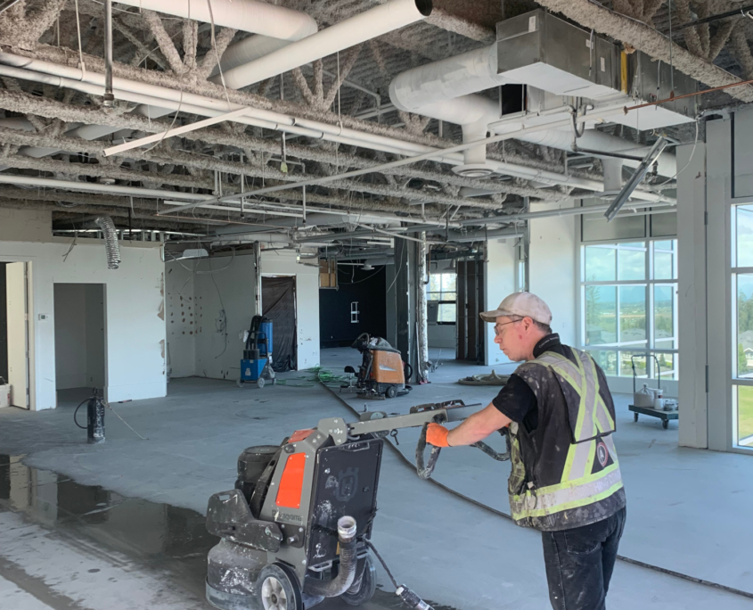 polish-concrete-4 A construction worker in a safety vest and helmet operates a floor grinding machine, preparing the surface for polished concrete inside a large, unfinished building with exposed pipes and ducts. Sunlight streams through the spacious windows.