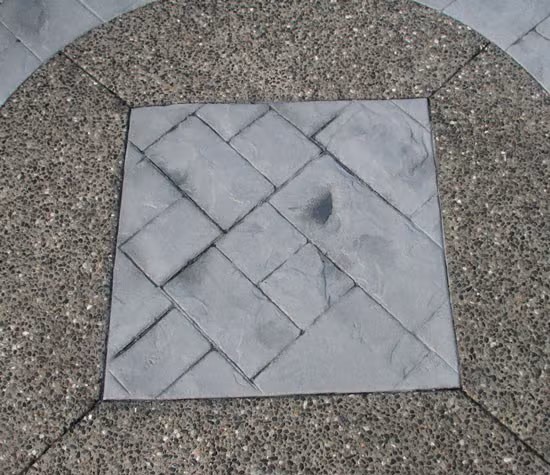 A square section of smooth, antiquing stamped concrete with a tile pattern is set within rough, exposed aggregate concrete forming a circular border around it.