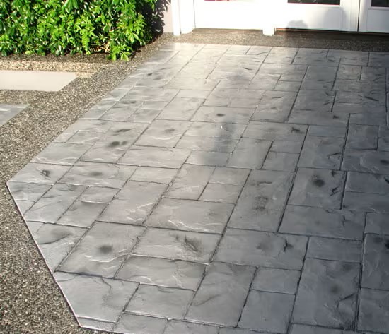 Stamped concrete patio with a textured, gray stone pattern featuring antiquing stamped concrete, bordered by exposed aggregate concrete. Green bushes are visible in the upper left corner near white-framed glass doors.