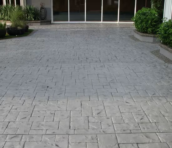 A concrete driveway with an antiquing stamped concrete, textured surface extends toward a building with large glass sliding doors. Green shrubs and potted plants line the sides of the entryway.