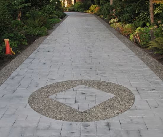 A long, paved driveway with an antiquing stamped concrete circular design near the center, featuring a diamond shape in the middle. The driveway is bordered by plants and trees, with orange cones on the left side.