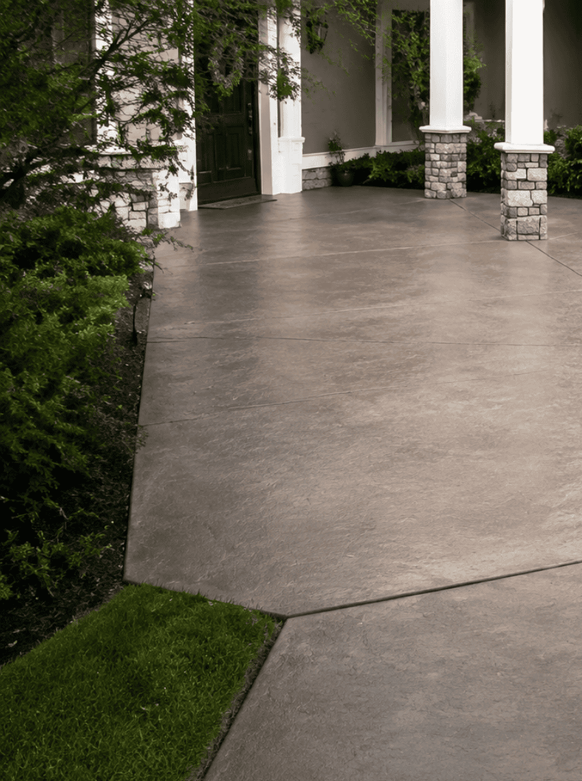 cr-con-2 Smooth concrete driveway—recently enhanced with expert cracked concrete repair—leads to a house with white columns and stone bases, surrounded by neatly trimmed green bushes and landscaped greenery.