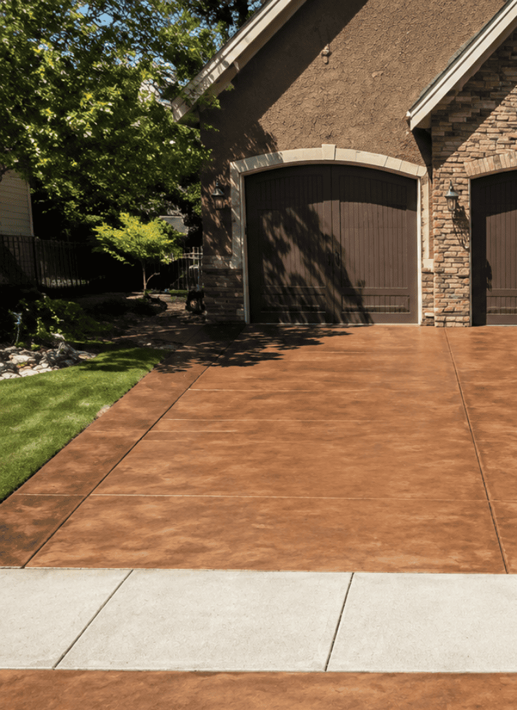 cr-con-3 A house with two brown garage doors, a wide brown concrete driveway featuring expertly done cracked concrete repair, a matching sidewalk, and landscaped shrubs and plants along the sides.