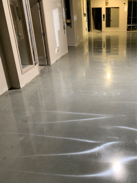 polish-con A hallway with polished concrete floors reflecting light. The sleek polished concrete surface contrasts with the unfinished walls, which feature visible patches and open electrical outlets.