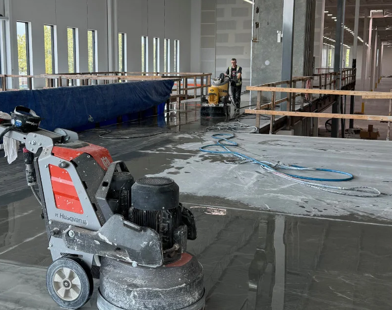 A concrete floor polishing machine sits on a shiny, wet surface in a large industrial building under construction, with workers and equipment visible in the background.