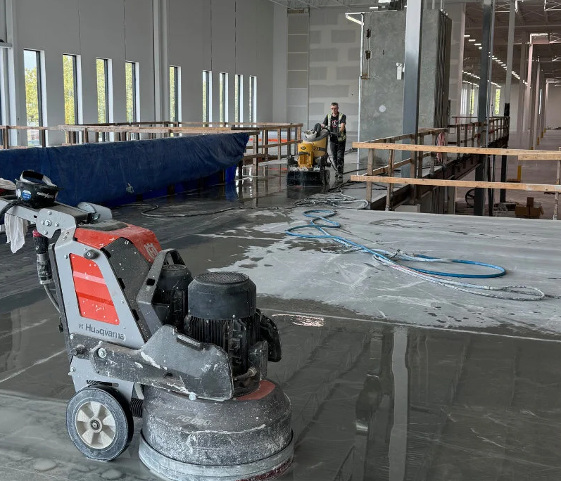 A concrete floor polishing machine sits on a shiny, wet surface in a large industrial building under construction, with workers and equipment visible in the background.