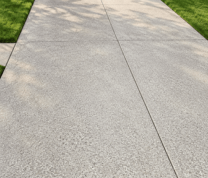 A clean, light gray concrete driveway—recently maintained to repair concrete cracks—leads up to a closed garage door, bordered by green grass lawns and parts of two suburban houses.
