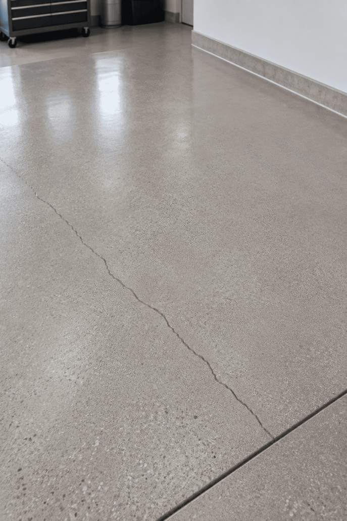 A polished concrete floor with a long, visible crack running across it, suggesting the need to repair concrete in this area. There are some reflections on the surface and a tool chest is partially visible in the background.