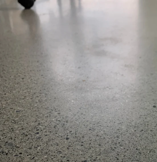 A close-up view of a polished concrete floor in an office, with rolling chairs in the background and large windows letting in natural light that highlights the smooth, reflective surface.